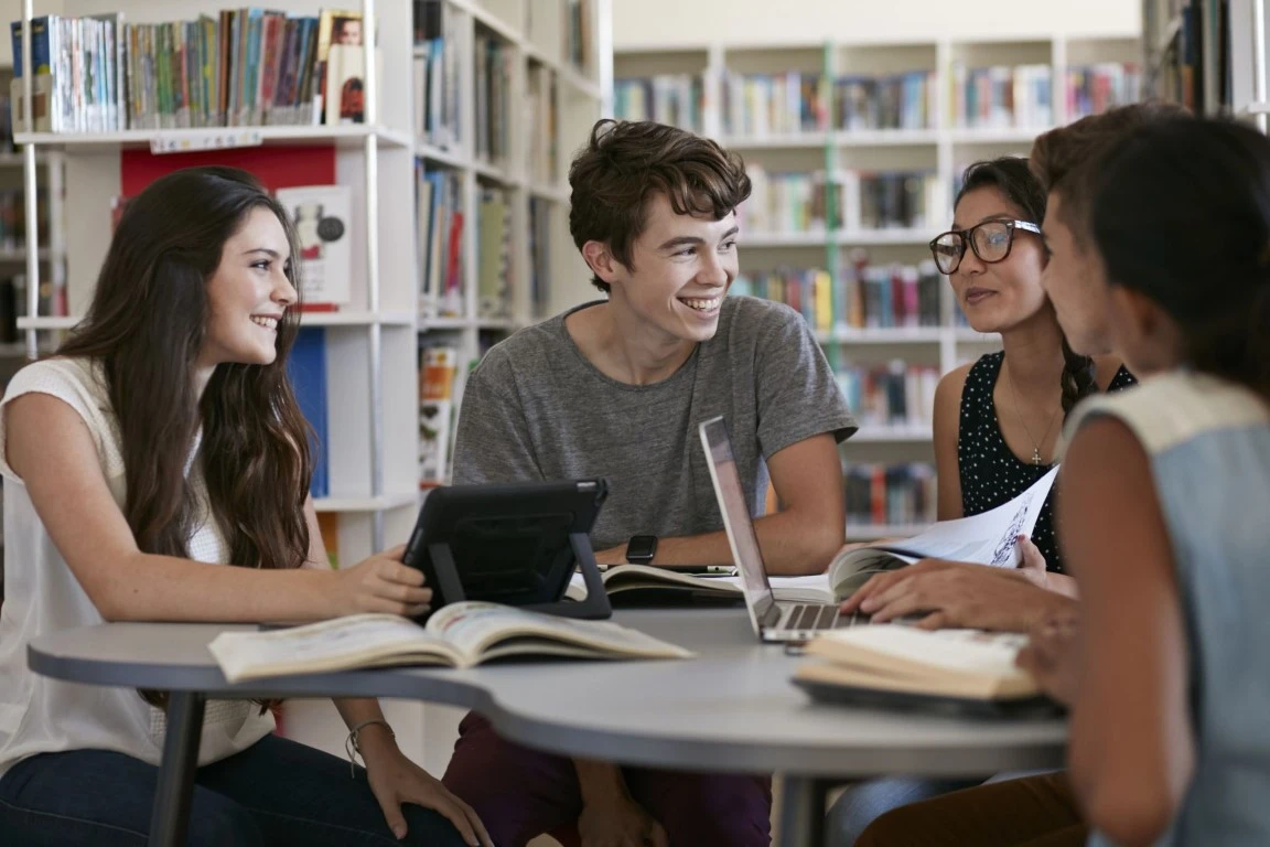 Grupa studentów podczas wspólnej nauki w bibliotece, pracujących z laptopami i tabletami przy okrągłym stole, w tle widoczne regały z książkami