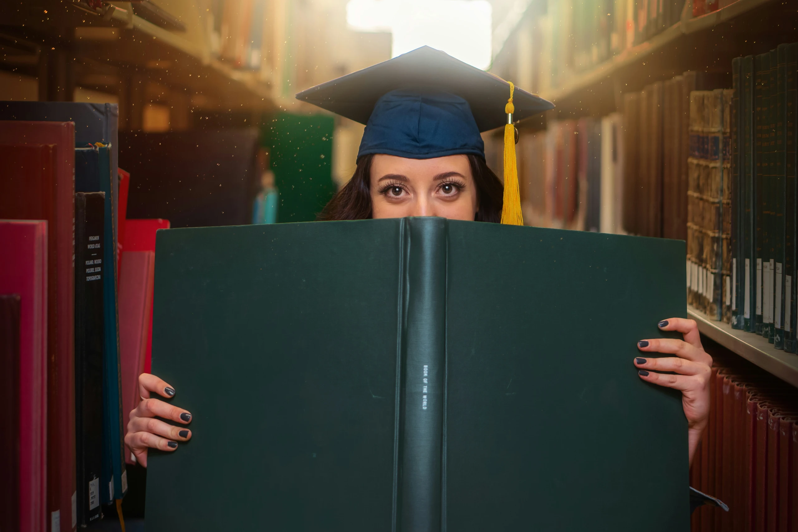 Kreatywne zdjęcie absolwentki w birecie akademickim, trzymającej dużą zieloną księgę zasłaniającą połowę twarzy. W tle widoczne regały biblioteczne i efekt świetlny.