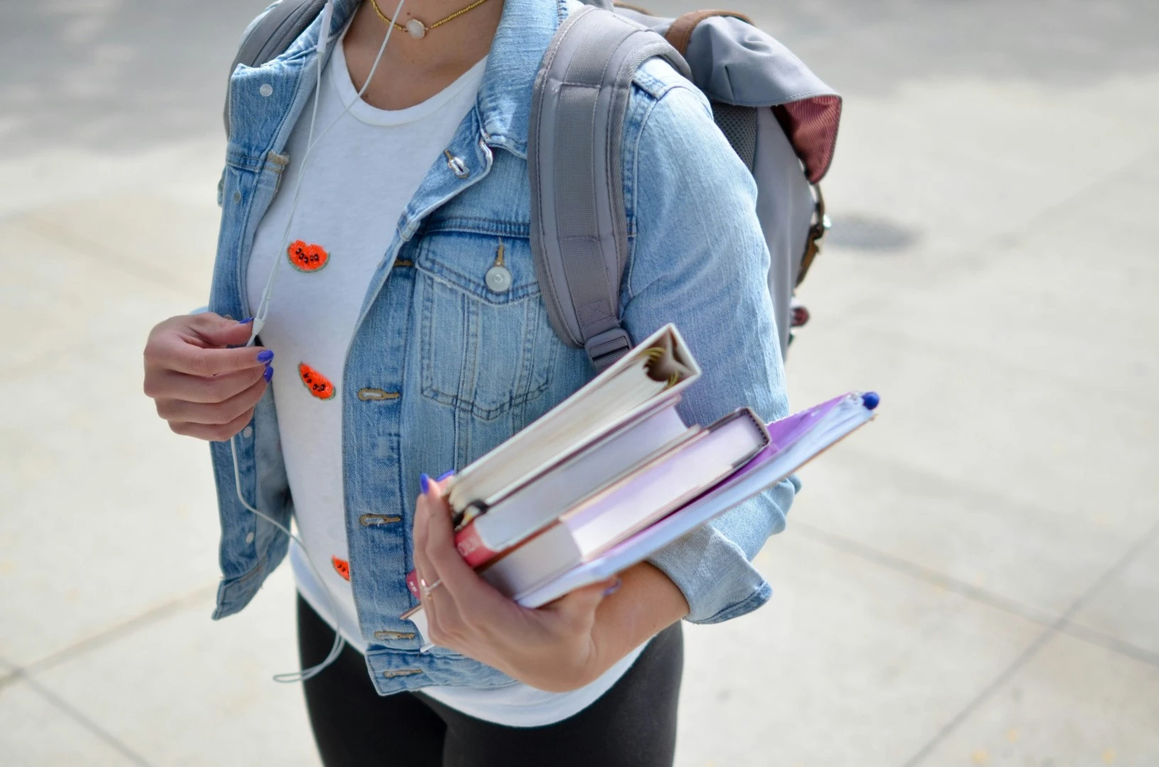 Zbliżenie na studenta w jasnej koszulce i jeansowej kurtce, trzymającego kilka podręczników. Widoczne są niebieskie paznokcie, biały t-shirt z pomarańczowymi przypinkami oraz szary plecak na ramieniu.