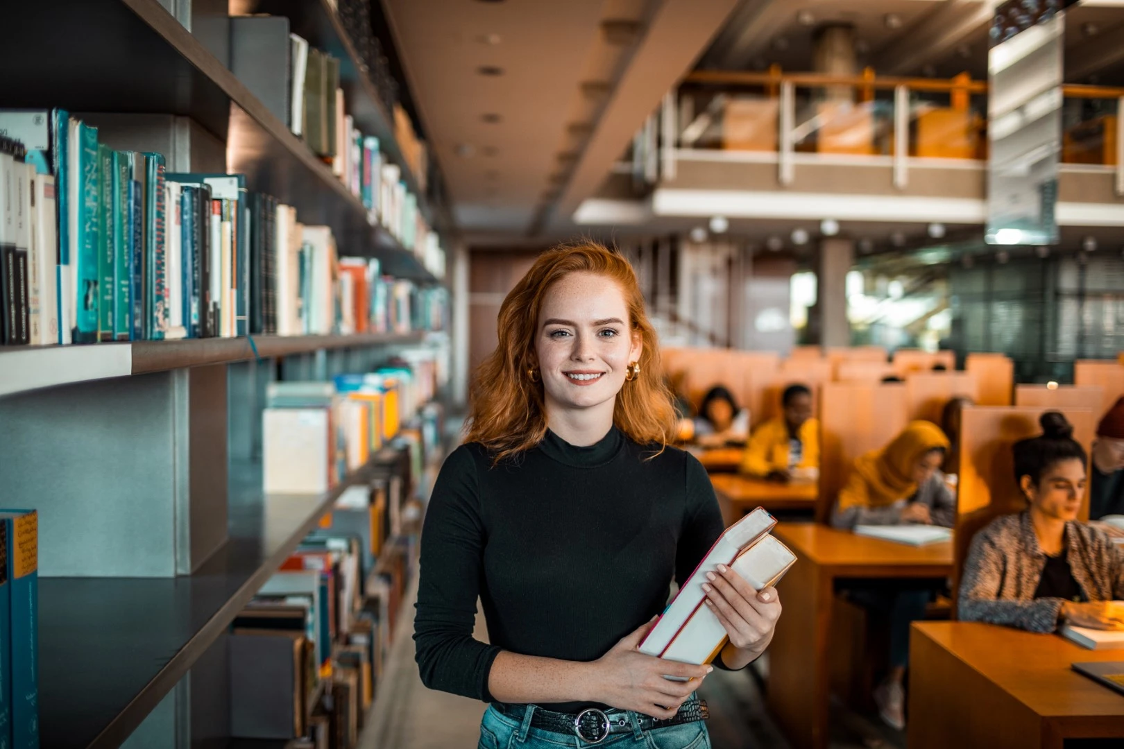 Uśmiechnięta studentka w czarnym golfie stojąca w bibliotece uniwersyteckiej, trzymająca książki. W tle widoczne regały z książkami oraz inni studenci uczący się przy stolikach.